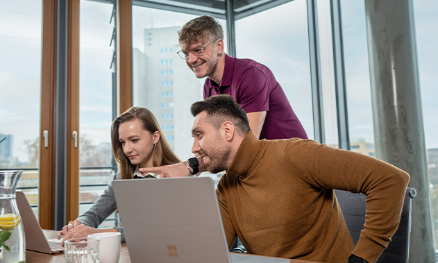Drei Personen befinden sich in einem Büroraum und schauen lächelnd auf einen Laptop.