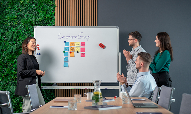 Vier Personen befinden sich in einem Büro. Drei Personen klatschen, während eine Person vor einem Whiteboard steht und etwas präsentiert.