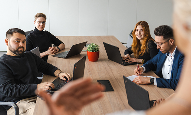 Vier Personen sitzen mit Ihren Laptops an einem Meeting Tisch. Die Perspektive des Bildes zeigt eine Person die vor den anderen Präsentiert.