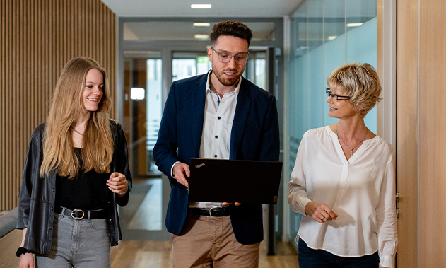 Drei Personen gehen über einen Flur. Die Person in der Mitte hält einen Laptop.